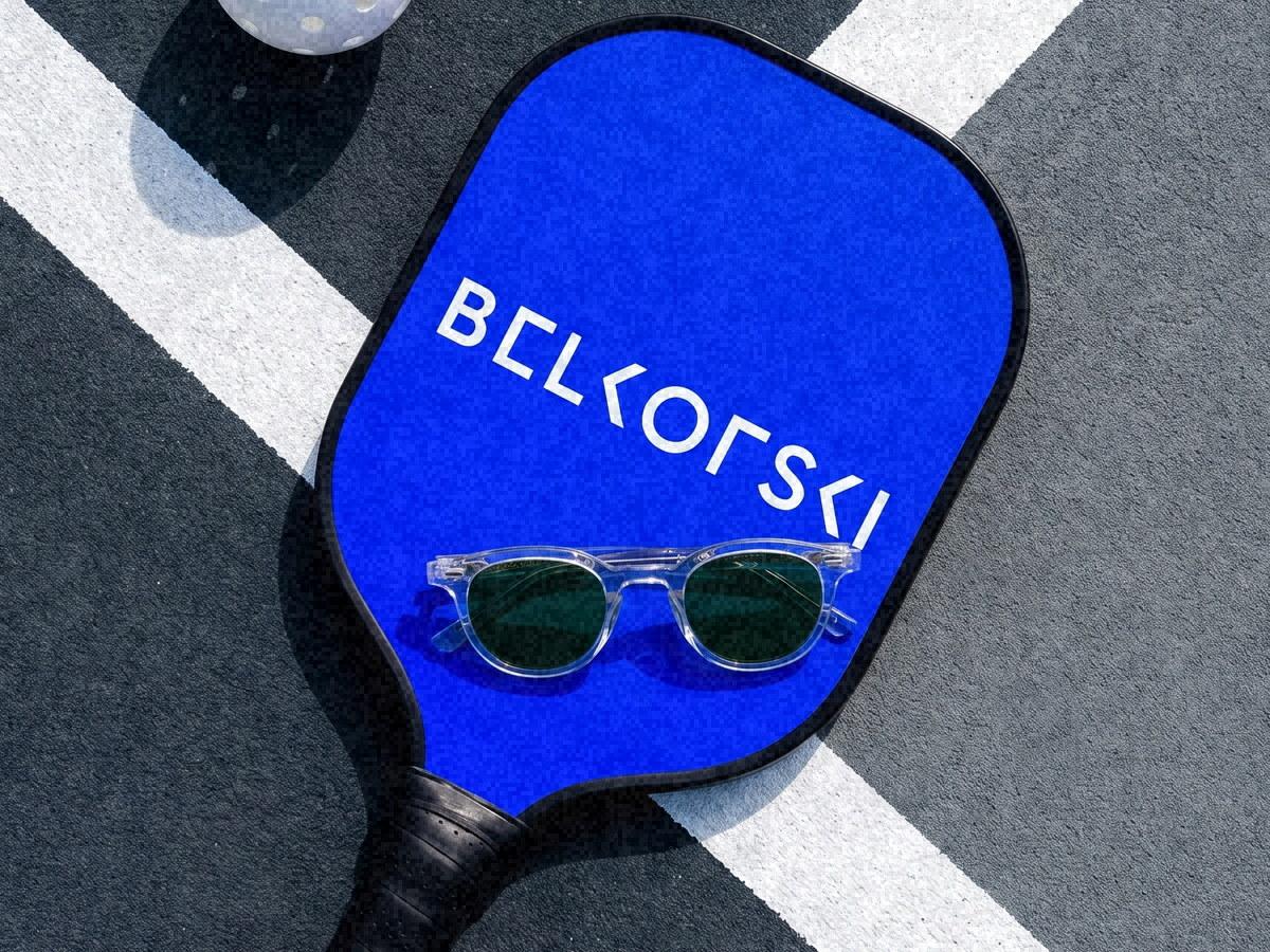 A blue Belkofski paddle and a pair of clear frames on a court, cut by the white line, shot from above.