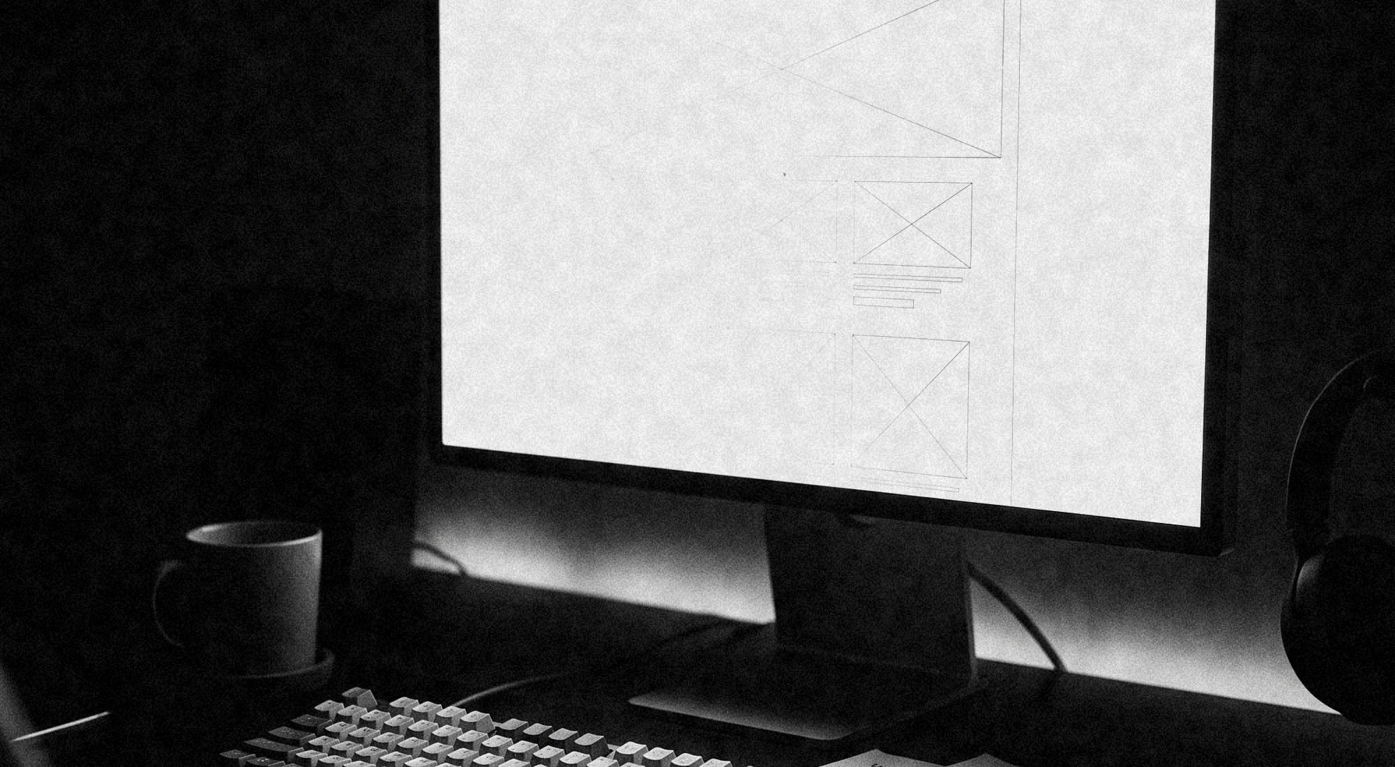A desk at night in black and white: a monitor showing a wireframe layout, a keyboard and wireframe sketches on paper beside it.