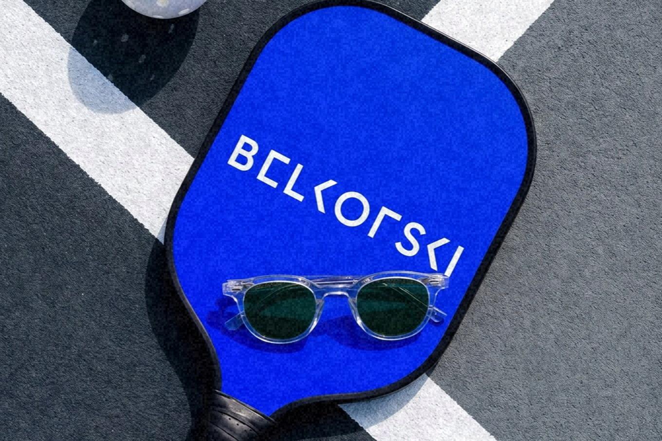 A blue Belkofski paddle carrying the wordmark, lying across a court line with a white ball beside it and a pair of clear frames on its face, shot from above.