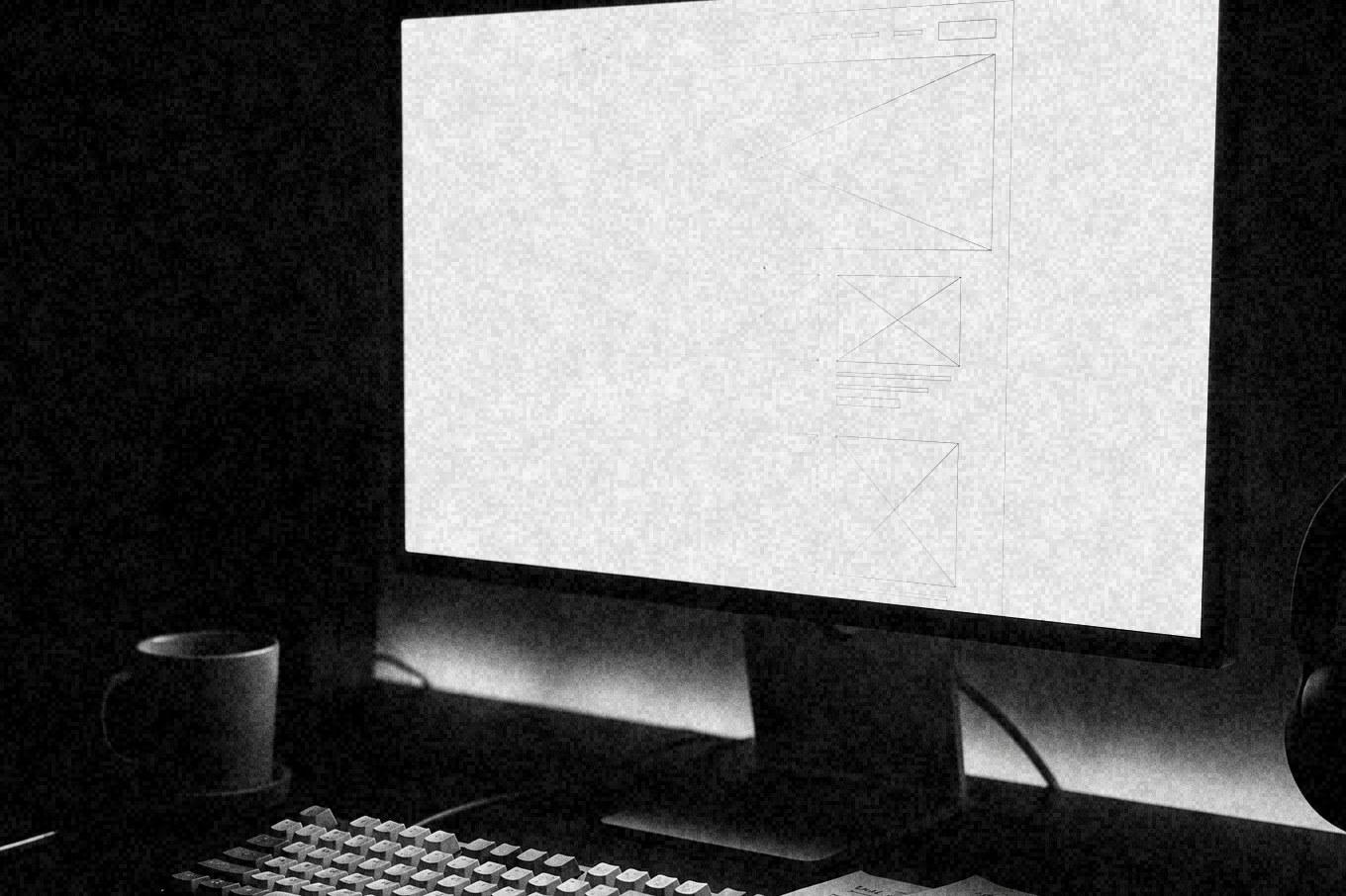 A desk at night in black and white: a monitor showing a wireframe layout, a keyboard and wireframe sketches on paper.